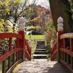 The red-painted wooden Japanese Bridge at Clyne Gardens with white finials on its posts, looking across the deck to stone steps and autumn trees