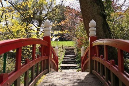 The red-painted wooden Japanese Bridge at Clyne Gardens with white finials on its posts, looking across the deck to stone steps and autumn trees