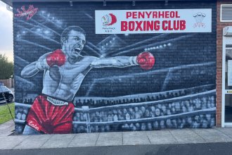 Black and white mural of boxer Colin Jones throwing a punch in red gloves and red shorts, on the wall of Penyrheol Boxing Club beneath the club sign.