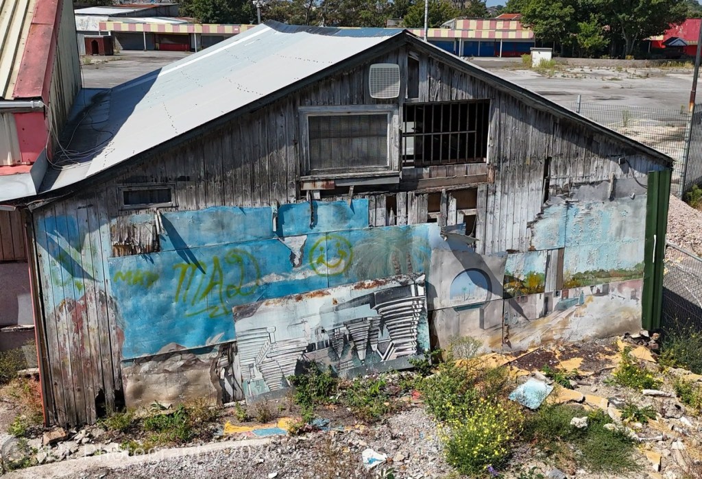 A weathered timber building with a faded, graffitied mural and broken boards, among weeds and rubble, with the empty funfair site and its striped stall fronts behind