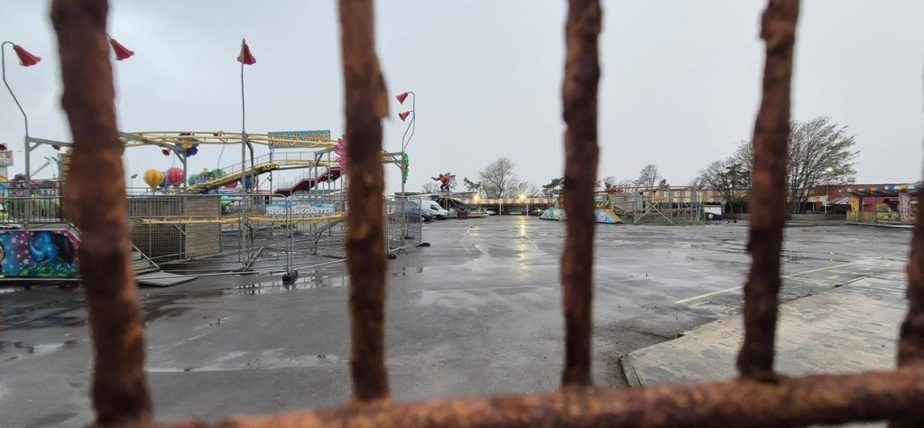Idle fairground rides, including a yellow rollercoaster, on an empty, rain-soaked Coney Beach site, seen through rusty railings