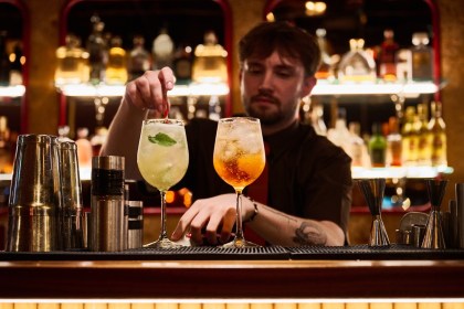 A bartender garnishes two cocktails on a dark bar in front of a lit back bar of bottles at a Cosy Club venue