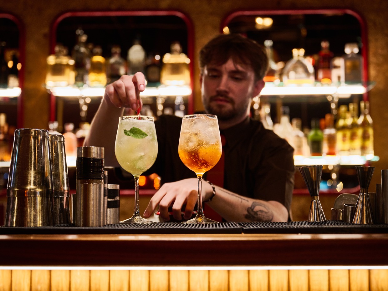A bartender garnishes two cocktails on a dark bar in front of a lit back bar of bottles at a Cosy Club venue