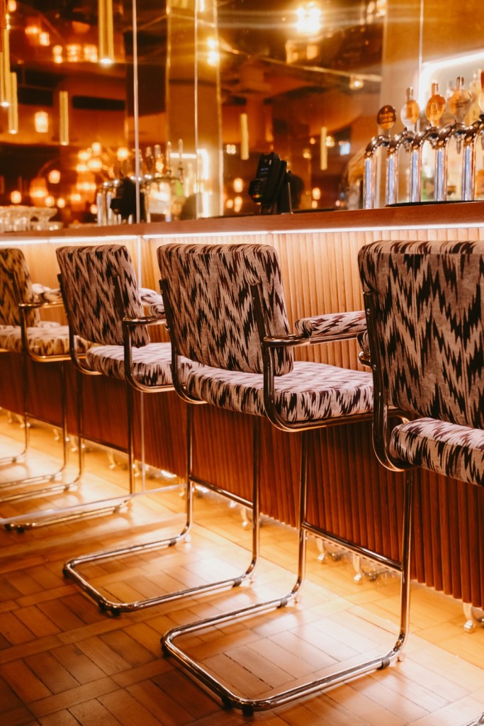 Patterned chrome-framed bar stools along a fluted wooden bar with a lit back bar behind at a Cosy Club