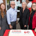Four people standing behind a large iced cake reading Cross Engineering, Celebrating 60 Years in Business, outside the firm’s entrance decorated with red, black and white balloons