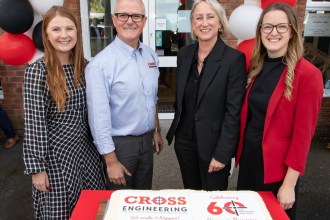 Four people standing behind a large iced cake reading Cross Engineering, Celebrating 60 Years in Business, outside the firm’s entrance decorated with red, black and white balloons