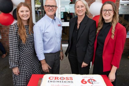 Four people standing behind a large iced cake reading Cross Engineering, Celebrating 60 Years in Business, outside the firm’s entrance decorated with red, black and white balloons