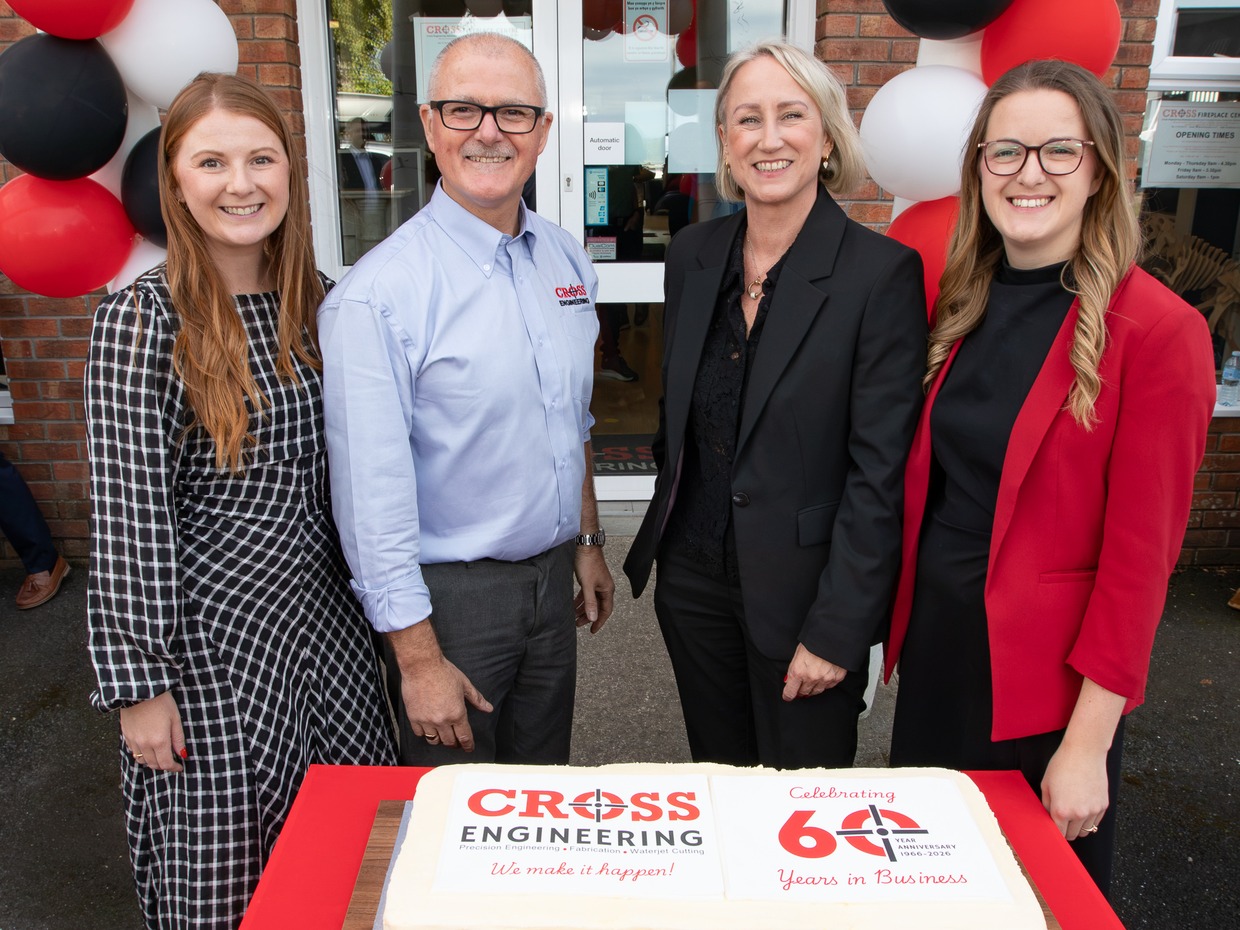 Four people standing behind a large iced cake reading Cross Engineering, Celebrating 60 Years in Business, outside the firm’s entrance decorated with red, black and white balloons