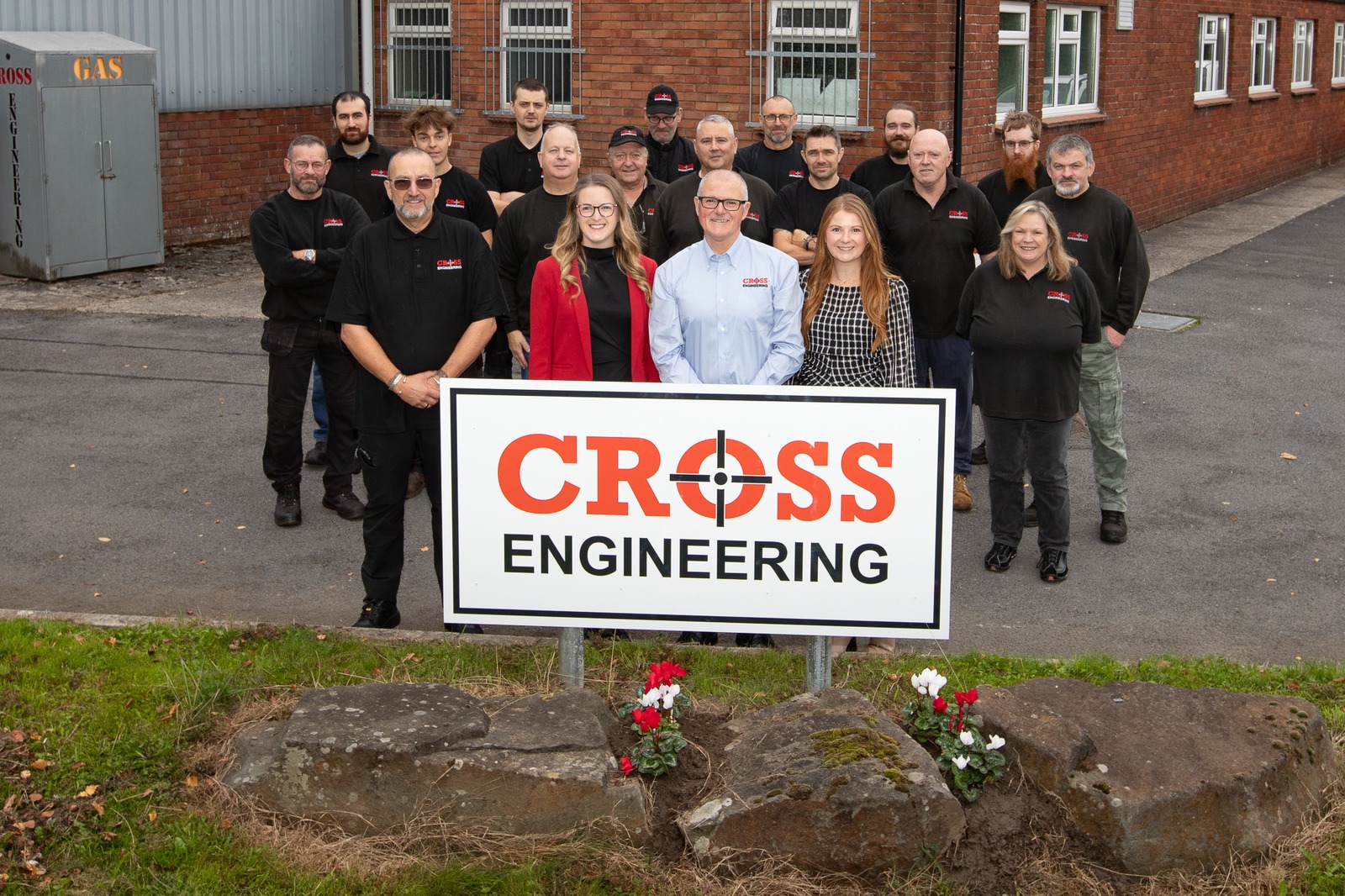 Around 20 Cross Engineering staff, most in black company polo shirts, standing behind a red and white Cross Engineering sign on a grass bank