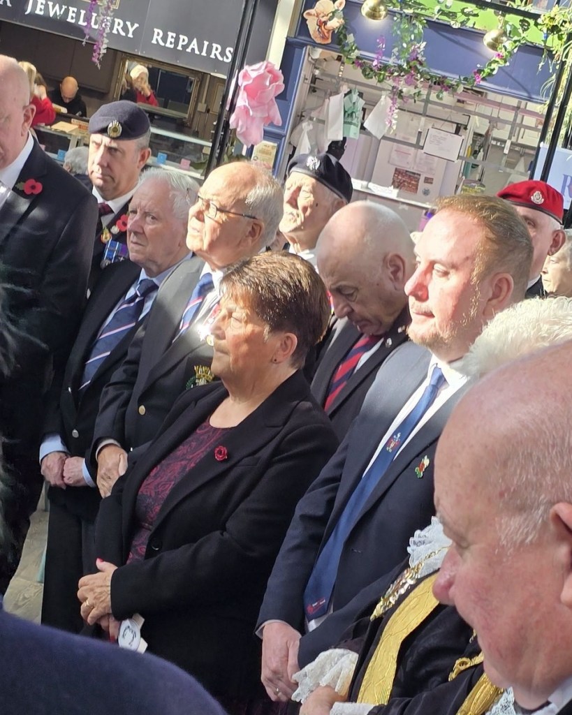 Veterans, councillors and members of the public stand in silence among the stalls of Swansea Market, several wearing poppies and berets.
