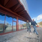 Two people walking along Cupid Way past the glazed shopfronts beneath the copper-coloured canopy of Copr Bay North car park on a sunny day