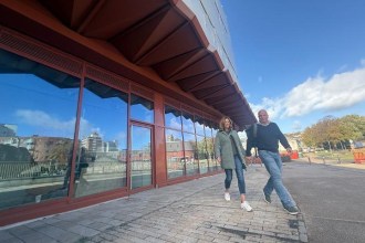 Two people walking along Cupid Way past the glazed shopfronts beneath the copper-coloured canopy of Copr Bay North car park on a sunny day