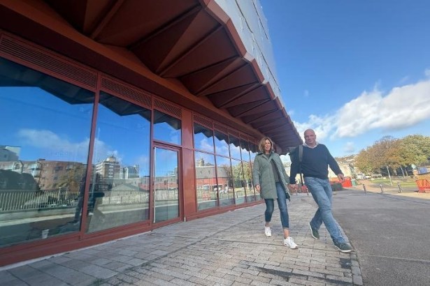 Two people walking along Cupid Way past the glazed shopfronts beneath the copper-coloured canopy of Copr Bay North car park on a sunny day