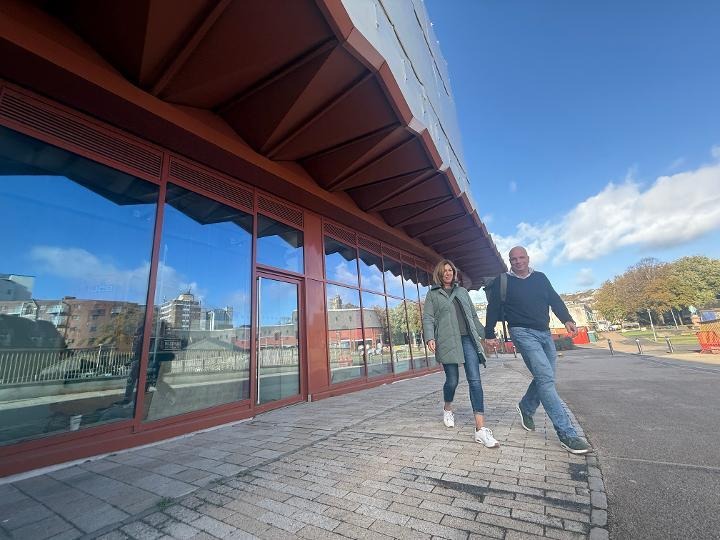 Two people walking along Cupid Way past the glazed shopfronts beneath the copper-coloured canopy of Copr Bay North car park on a sunny day