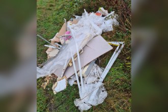 A pile of fly-tipped plasterboard sheets, plastic sheeting, white uPVC trim and metal beading on a grass verge beside brambles