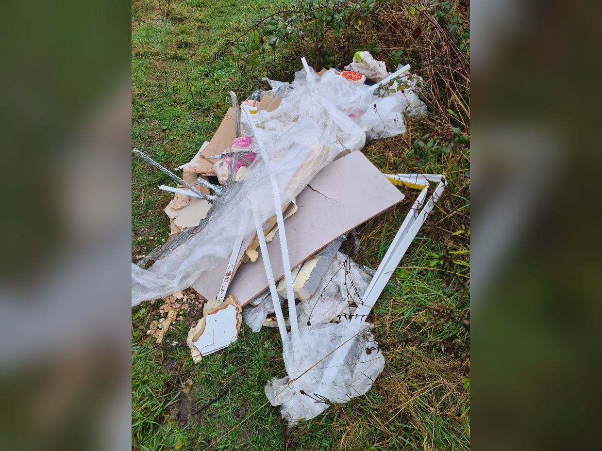 A pile of fly-tipped plasterboard sheets, plastic sheeting, white uPVC trim and metal beading on a grass verge beside brambles