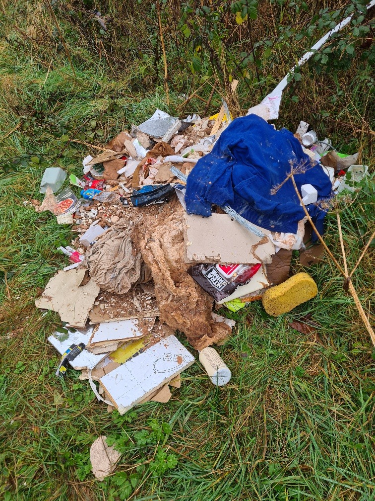 A second pile of fly-tipped waste on grass, with broken plasterboard, plaster rubble, insulation, a blue cloth and empty tubes and packaging