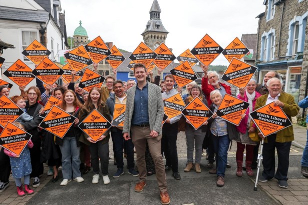 David Chadwick in a checked jacket smiling in front of a crowd of supporters holding orange Welsh Liberal Democrats diamond placards