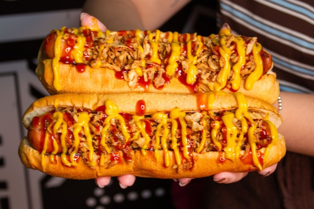 Two hands holding two long hot dogs covered in crispy onions, ketchup and mustard