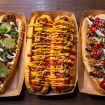 Three long hot dogs in cardboard trays seen from above, one with pickles and spring onion, one with crispy onions, ketchup and mustard, one with red chilli and sesame seeds