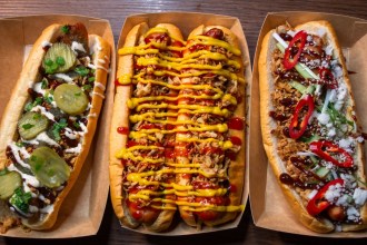 Three long hot dogs in cardboard trays seen from above, one with pickles and spring onion, one with crispy onions, ketchup and mustard, one with red chilli and sesame seeds