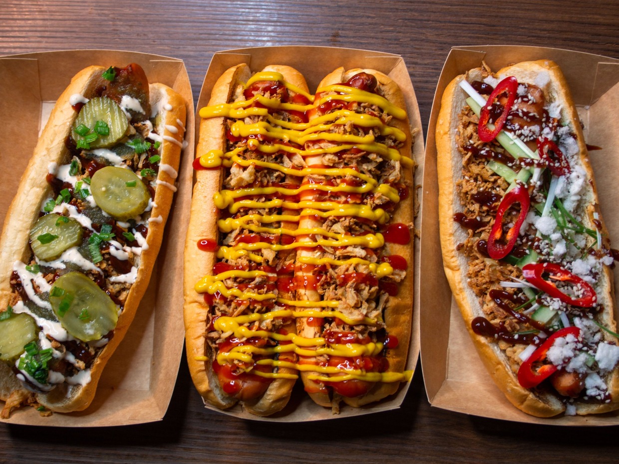 Three long hot dogs in cardboard trays seen from above, one with pickles and spring onion, one with crispy onions, ketchup and mustard, one with red chilli and sesame seeds