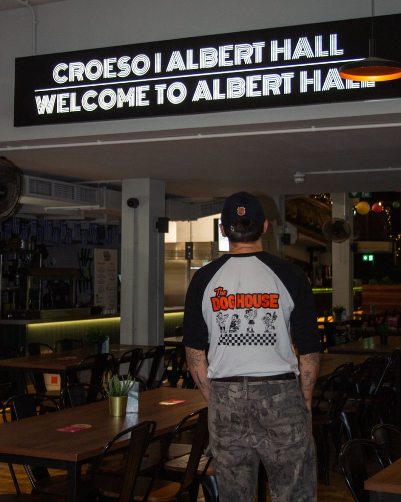 A man seen from behind in a Dog House baseball shirt stands under an illuminated sign reading Croeso i Albert Hall, Welcome to Albert Hall, above rows of tables