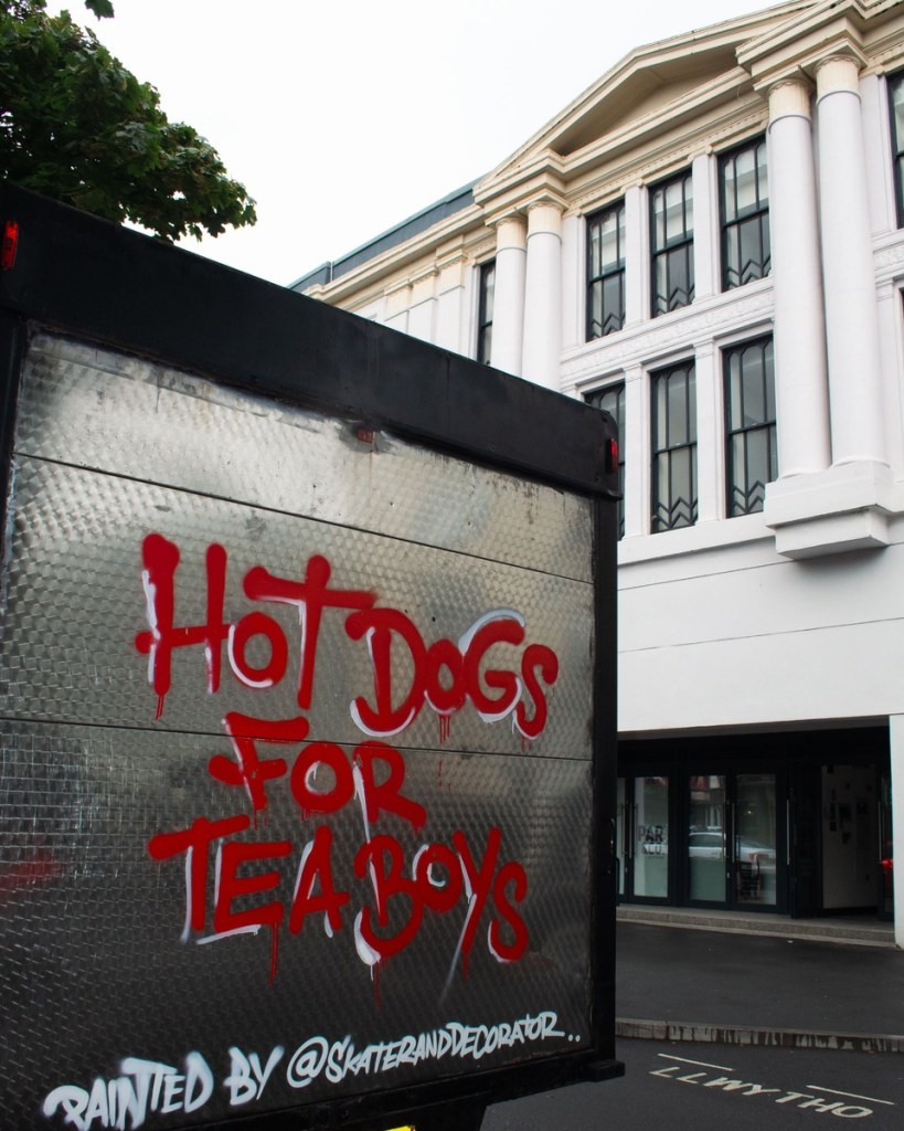 The back door of the truck with Hot Dogs for Tea Boys spray-painted in dripping red letters, signed by Skater and Decorator, beside a white columned building
