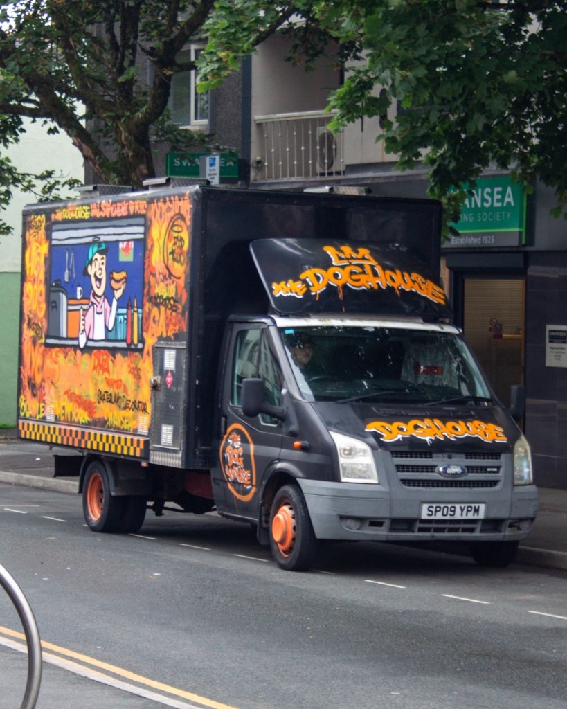 A black food truck spray-painted in orange and yellow with The Dog House on the bonnet and a cartoon hot dog vendor on the side, parked on a Swansea street