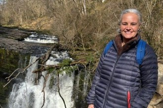Eleanor Barnfield smiling in a navy puffer jacket and blue rucksack on a rocky path beside a waterfall