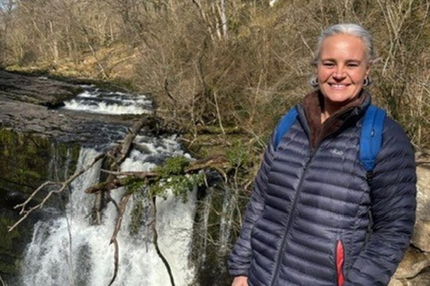 Eleanor Barnfield smiling in a navy puffer jacket and blue rucksack on a rocky path beside a waterfall