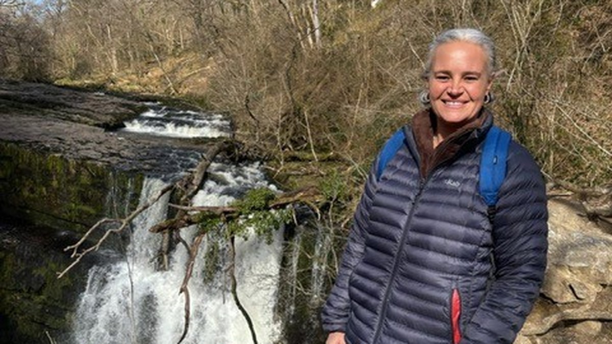 Eleanor Barnfield smiling in a navy puffer jacket and blue rucksack on a rocky path beside a waterfall