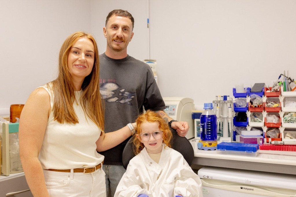 Esme Mariani in a white lab coat and glasses, sitting in front of her parents Emily and Mathew beside a laboratory bench