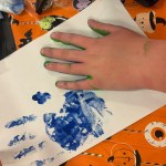 A child's hand with green paint on the fingertips resting on white paper next to a blue painted handprint, on an orange Halloween tablecloth