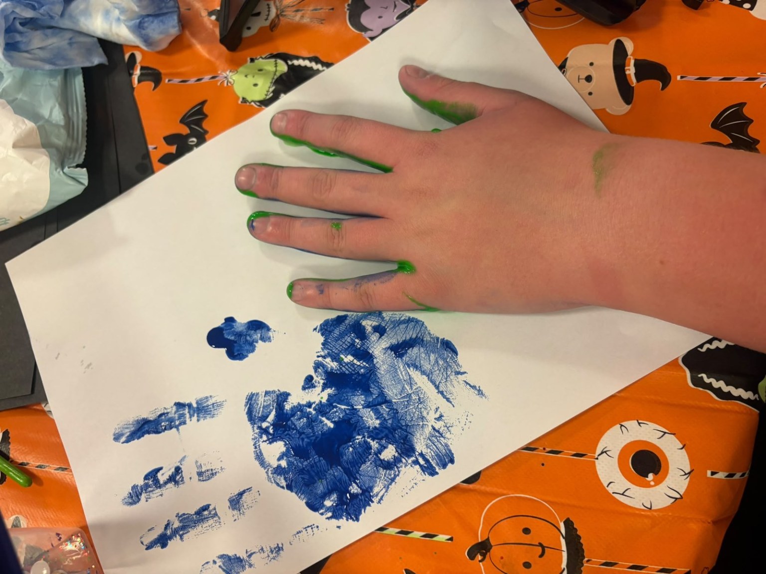 A child's hand with green paint on the fingertips resting on white paper next to a blue painted handprint, on an orange Halloween tablecloth