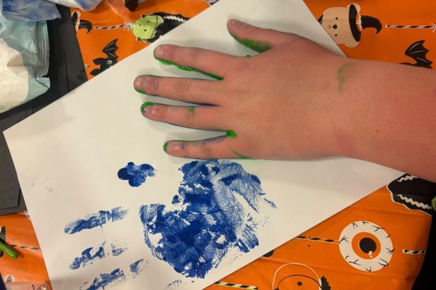 A child's hand with green paint on the fingertips resting on white paper next to a blue painted handprint, on an orange Halloween tablecloth