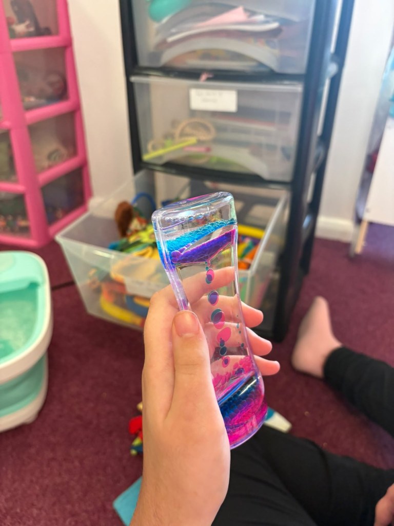 A child's hand holding a sensory liquid timer with pink and blue bubbles in a playroom with storage drawers and toy boxes behind