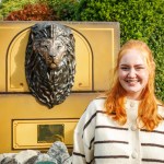 A smiling young woman with long red hair in a striped cardigan beside a bronze lion head mounted on a gold-painted plinth