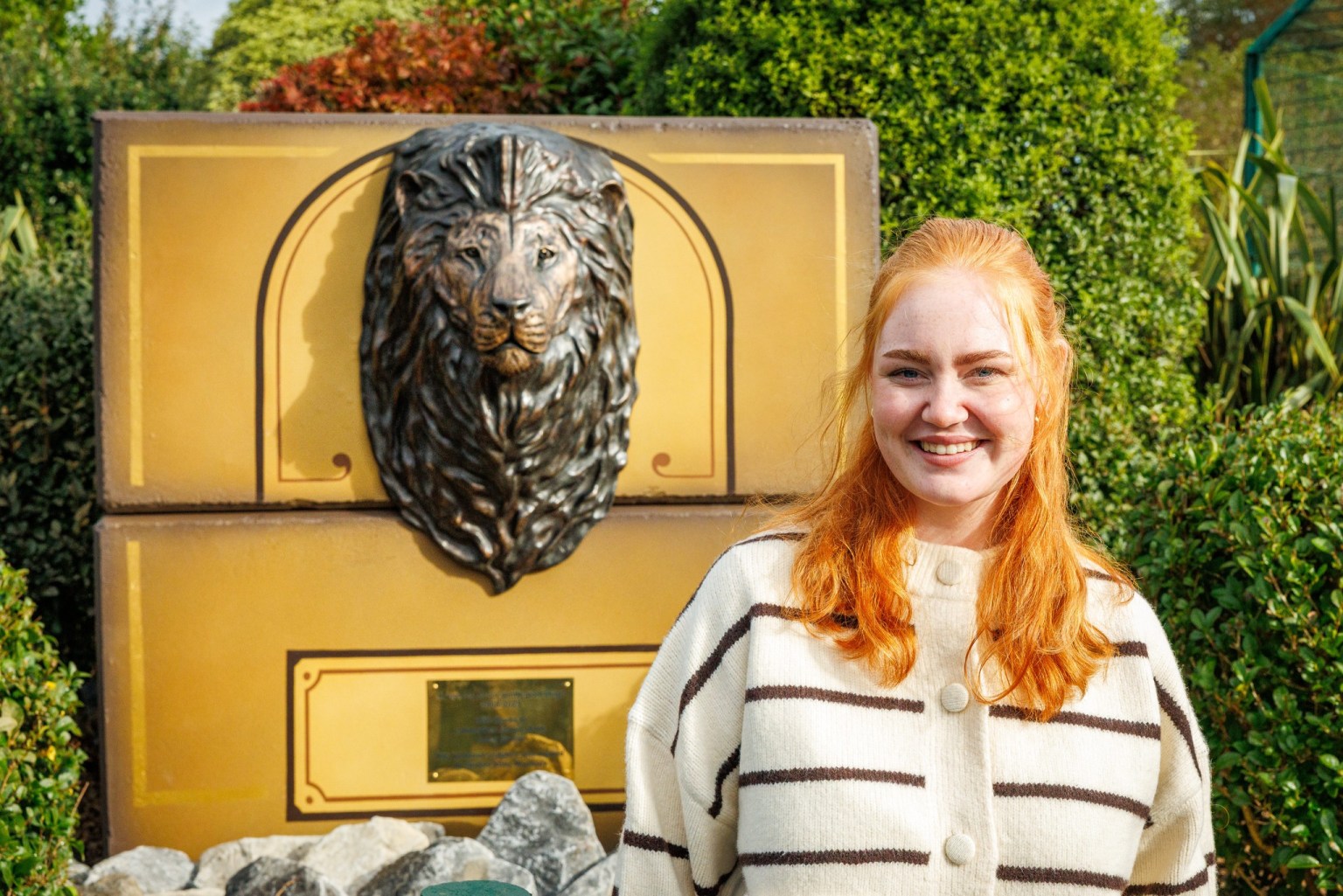 A smiling young woman with long red hair in a striped cardigan beside a bronze lion head mounted on a gold-painted plinth
