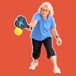 Gloria, a woman with white hair and glasses in a blue polo shirt and black cropped trousers, reaching to hit a yellow pickleball with a black and blue paddle, cut out against a coral background