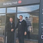 Two men shaking hands outside the new Gorseinon banking hub, with the Banking Hub and Post Office sign above the shopfront and a Banking Hub flag beside the door