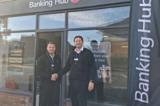 Two men shaking hands outside the new Gorseinon banking hub, with the Banking Hub and Post Office sign above the shopfront and a Banking Hub flag beside the door