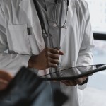 A doctor in a white coat with a stethoscope holds a tablet computer while a patient looks on