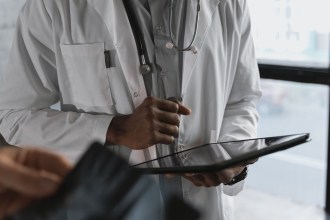 A doctor in a white coat with a stethoscope holds a tablet computer while a patient looks on