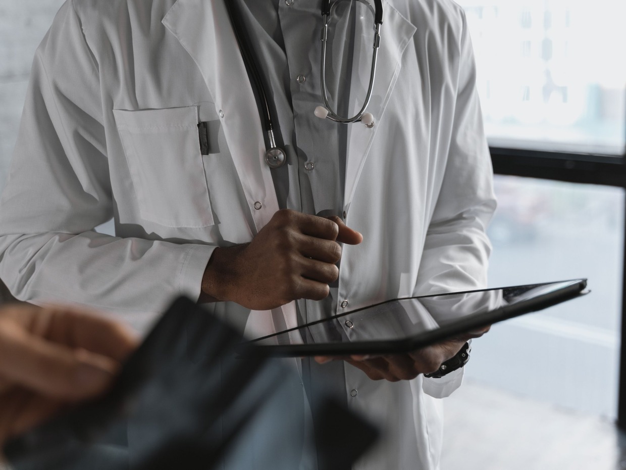 A doctor in a white coat with a stethoscope holds a tablet computer while a patient looks on