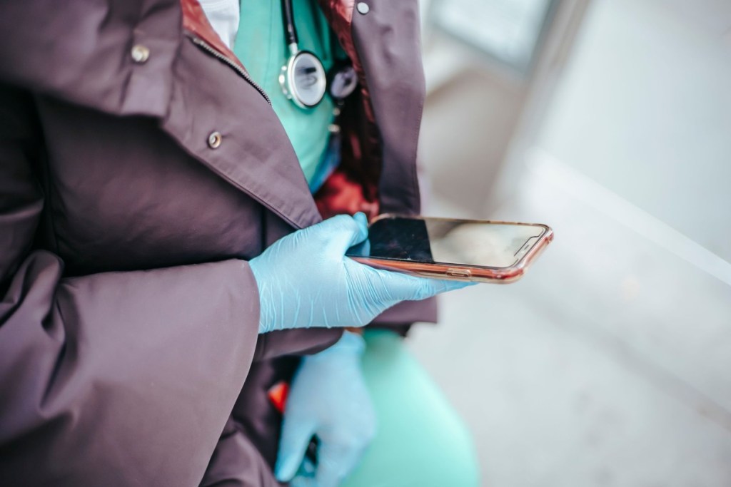 A healthcare worker in scrubs and blue gloves, with a stethoscope round their neck, holds a mobile phone