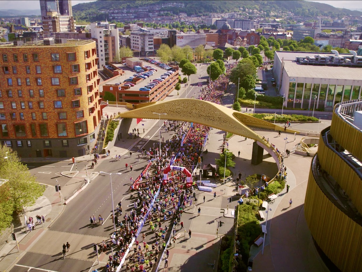 Aerial view of thousands of runners gathered at a start arch beneath the golden Copr Bay bridge in Swansea city centre