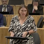 Heledd Fychan, in a black and white patterned dress, speaking at a lectern in the Senedd chamber with members seated behind her