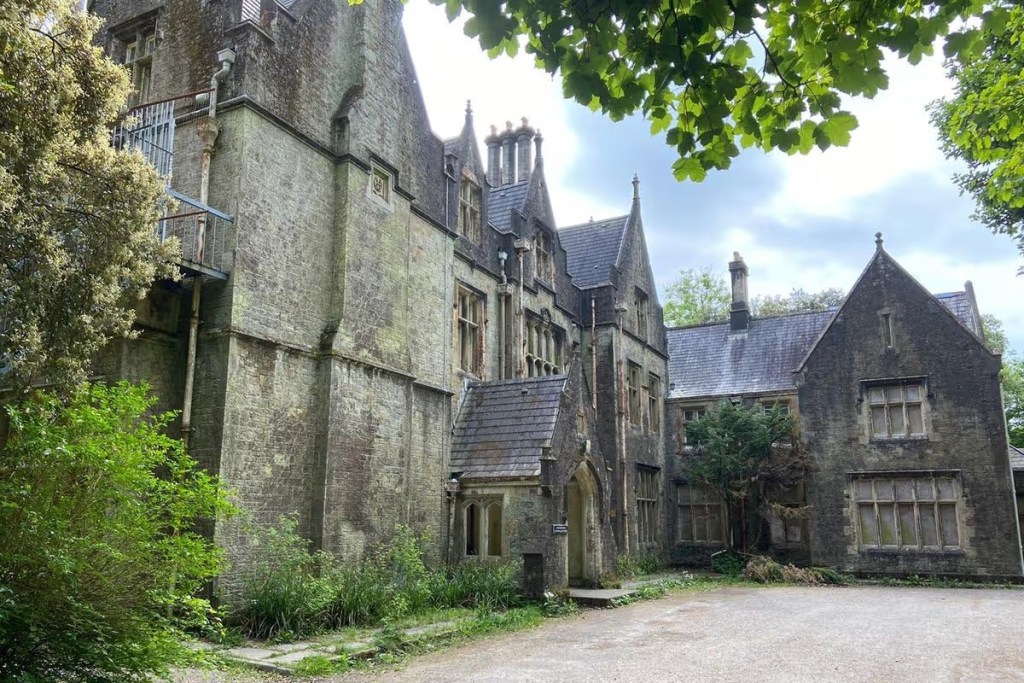 A large Tudor Gothic stone mansion with tall chimneys and steep gables, its walls streaked with moss and surrounded by overgrown trees