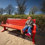 Esme Mariani, with red hair and glasses, smiling as she sits on a bright red British Heart Foundation bench on the sand at Swansea Beach